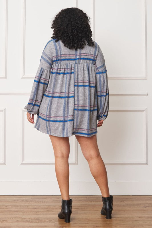 A person with curly hair wearing a Plus Plaid Empire Waist Balloon Sleeve Mini Dress stands against a white wall, smiling.