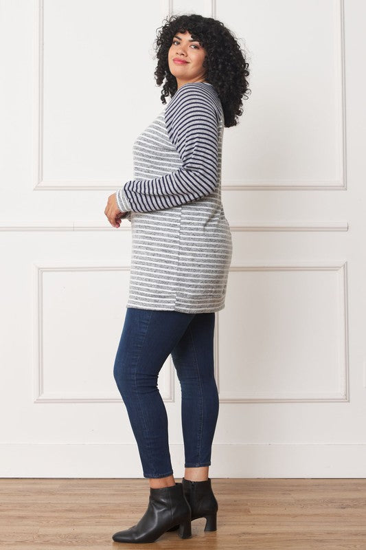 A woman stands smiling against a light-colored wall, showcasing a Plus Stripe Two Tone Tunic made of polyester spandex, paired with denim jeans.