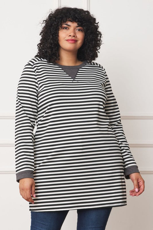 A person with curly hair smiles while wearing the Plus Stripe V-Stich Tunic, a black-and-white striped shirt designed in plus size, standing against a plain background.
