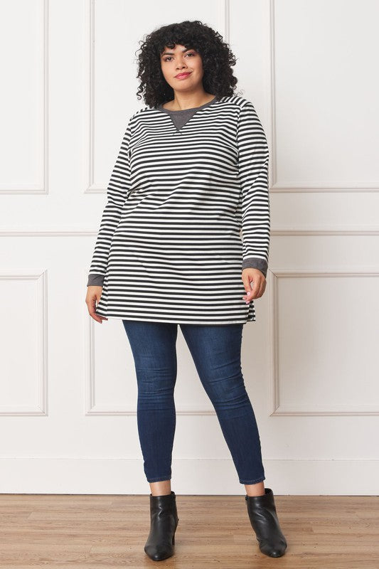 A person with curly hair smiles while wearing the Plus Stripe V-Stich Tunic, a black-and-white striped shirt designed in plus size, standing against a plain background.