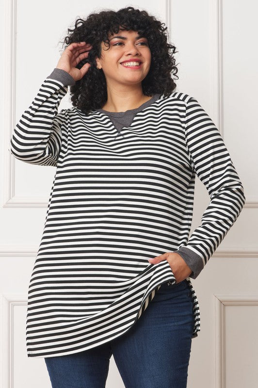A person with curly hair smiles while wearing the Plus Stripe V-Stich Tunic, a black-and-white striped shirt designed in plus size, standing against a plain background.