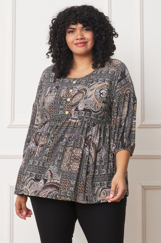 A person with curly hair wearing the Plus Button Accent Paisley Top and black pants stands in front of a white paneled wall, with one arm raised and the other resting by their side. Made in the United States.