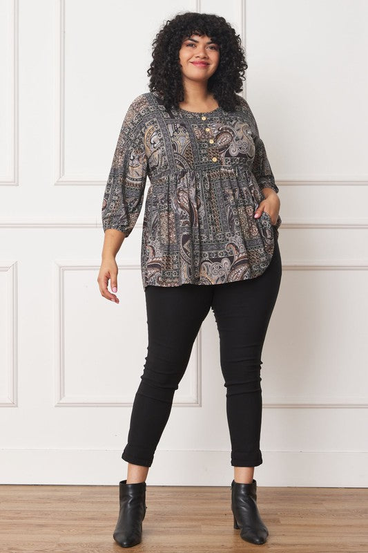 A person with curly black hair wears the Plus Button Accent Paisley Top paired with black pants, standing with one hand behind their head.