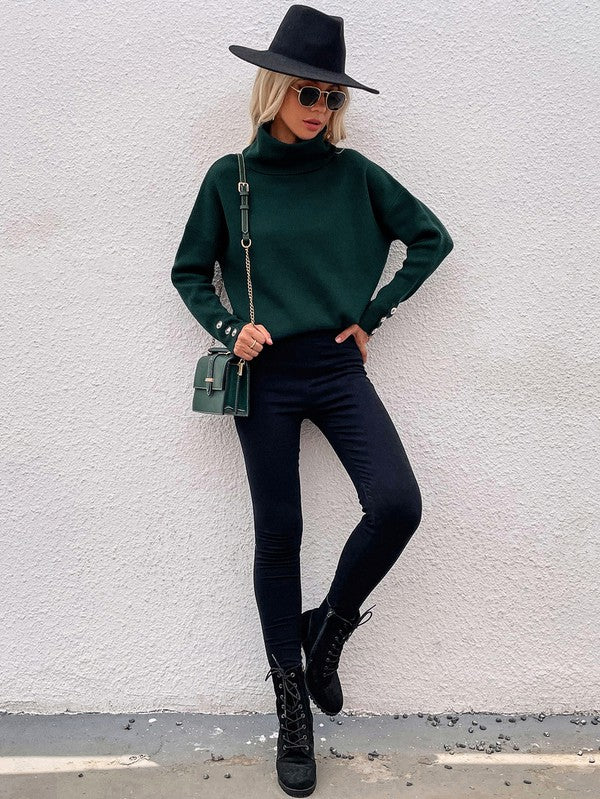 A woman modeling the Women's Long Sleeve Mock Neck Sweater in dark green, featuring button details on the sleeves, paired with black pants and posing against a textured white wall.