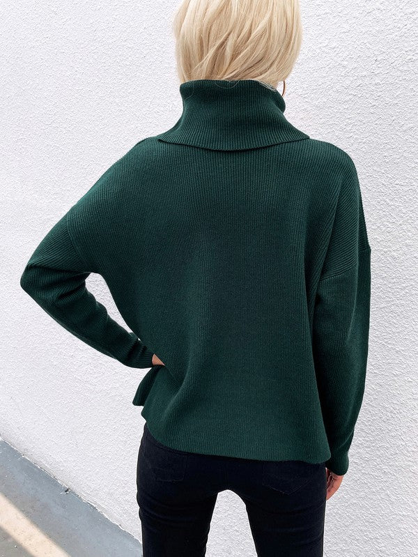 A woman modeling the Women's Long Sleeve Mock Neck Sweater in dark green, featuring button details on the sleeves, paired with black pants and posing against a textured white wall.