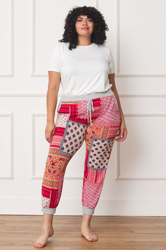A person is standing indoors, with only part of their upper body visible, wearing a white top and Plus Quilted Print Joggers featuring a colorful pattern. The joggers have a drawstring waist and pockets where the person's hands are tucked. Their left foot is bare.
