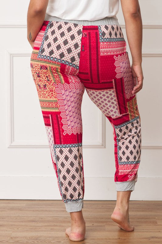 A person is standing indoors, with only part of their upper body visible, wearing a white top and Plus Quilted Print Joggers featuring a colorful pattern. The joggers have a drawstring waist and pockets where the person's hands are tucked. Their left foot is bare.