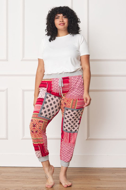 A person is standing indoors, with only part of their upper body visible, wearing a white top and Plus Quilted Print Joggers featuring a colorful pattern. The joggers have a drawstring waist and pockets where the person's hands are tucked. Their left foot is bare.