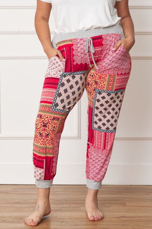 A person is standing indoors, with only part of their upper body visible, wearing a white top and Plus Quilted Print Joggers featuring a colorful pattern. The joggers have a drawstring waist and pockets where the person's hands are tucked. Their left foot is bare.