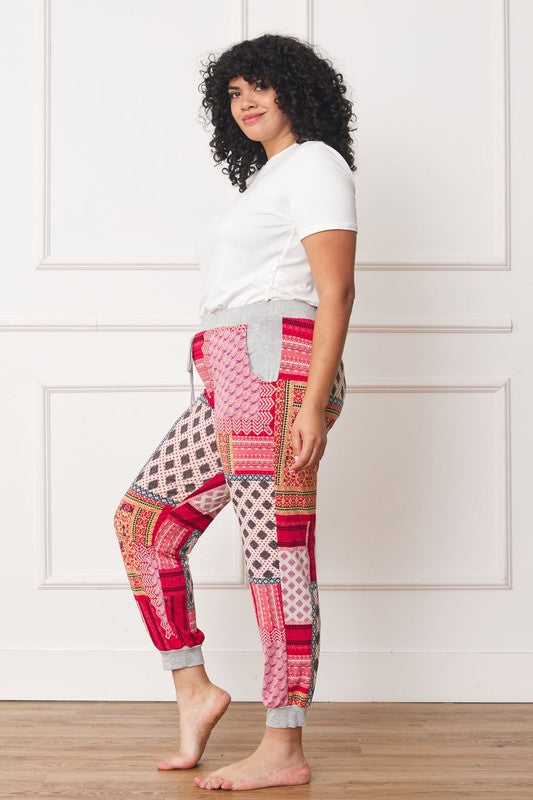A person is standing indoors, with only part of their upper body visible, wearing a white top and Plus Quilted Print Joggers featuring a colorful pattern. The joggers have a drawstring waist and pockets where the person's hands are tucked. Their left foot is bare.