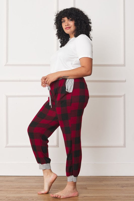 A person poses, wearing a white shirt and the stylish Plus Checkered Print Drawstring Jogger pants in red-and-black checkered pattern with gray cuffs and waistband, standing on a wooden floor against a white wall. These joggers feature convenient pockets and are made of 95% polyester for both comfort and style.