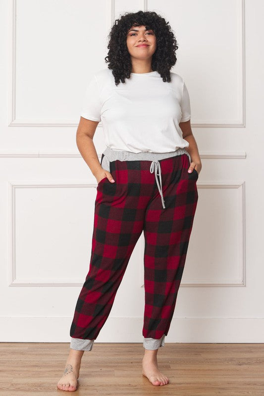 A person poses, wearing a white shirt and the stylish Plus Checkered Print Drawstring Jogger pants in red-and-black checkered pattern with gray cuffs and waistband, standing on a wooden floor against a white wall. These joggers feature convenient pockets and are made of 95% polyester for both comfort and style.
