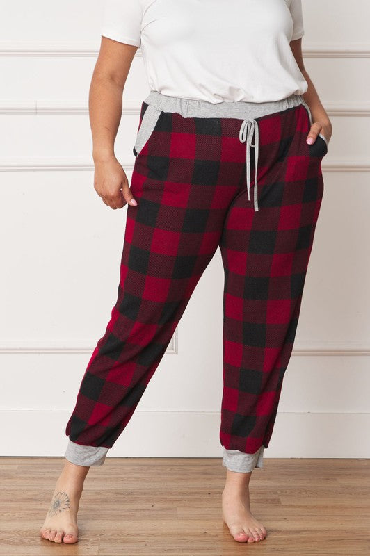 A person poses, wearing a white shirt and the stylish Plus Checkered Print Drawstring Jogger pants in red-and-black checkered pattern with gray cuffs and waistband, standing on a wooden floor against a white wall. These joggers feature convenient pockets and are made of 95% polyester for both comfort and style.