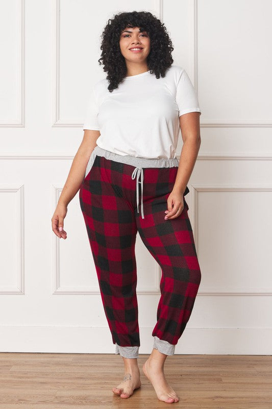 A person poses, wearing a white shirt and the stylish Plus Checkered Print Drawstring Jogger pants in red-and-black checkered pattern with gray cuffs and waistband, standing on a wooden floor against a white wall. These joggers feature convenient pockets and are made of 95% polyester for both comfort and style.