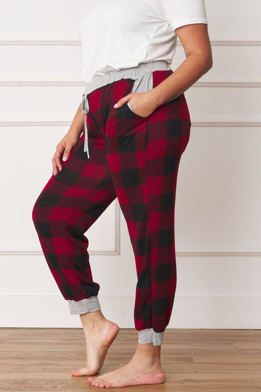 A person poses, wearing a white shirt and the stylish Plus Checkered Print Drawstring Jogger pants in red-and-black checkered pattern with gray cuffs and waistband, standing on a wooden floor against a white wall. These joggers feature convenient pockets and are made of 95% polyester for both comfort and style.