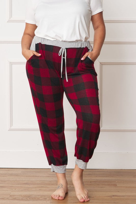 A person poses, wearing a white shirt and the stylish Plus Checkered Print Drawstring Jogger pants in red-and-black checkered pattern with gray cuffs and waistband, standing on a wooden floor against a white wall. These joggers feature convenient pockets and are made of 95% polyester for both comfort and style.