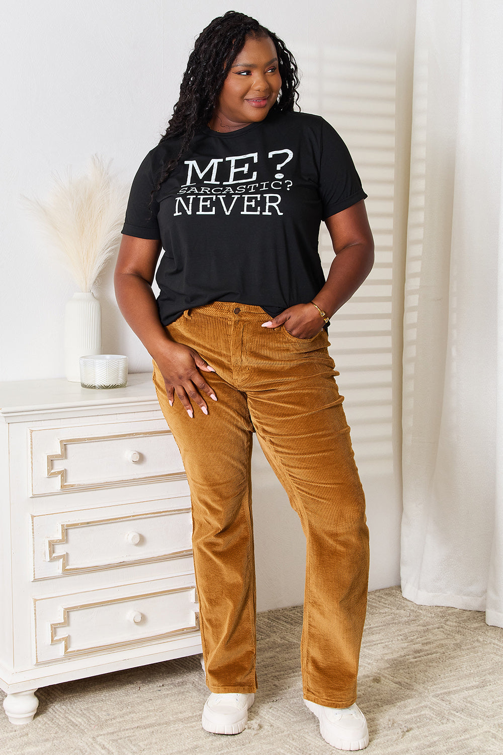 A woman with long dark hair smiles while wearing a trendy black Simply Love Letter Graphic Round Neck T-Shirt featuring the text "ME? Sarcastic? NEVER" and brown corduroy pants. She stands in a room with white walls and decor.