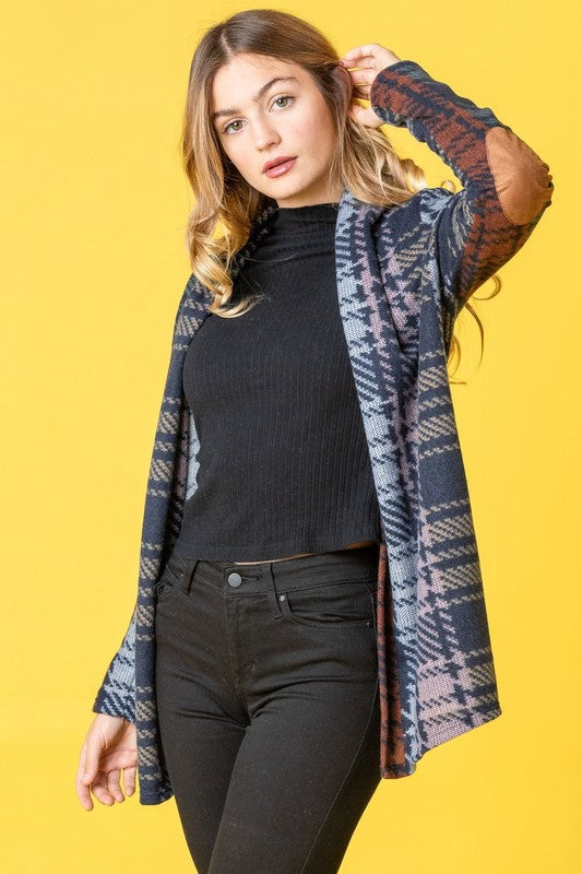 A woman wearing the Multi Color Plaid Elbow Patch Cardigan, made in the United States, strikes a pose with one hand on her cardigan and the other near her head, styled with a black top and black pants.