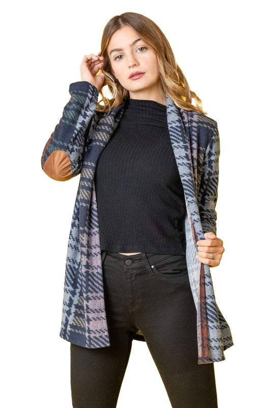 A woman wearing the Multi Color Plaid Elbow Patch Cardigan, made in the United States, strikes a pose with one hand on her cardigan and the other near her head, styled with a black top and black pants.