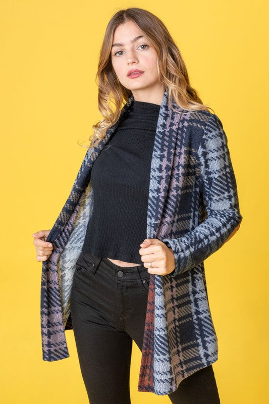 A woman wearing the Multi Color Plaid Elbow Patch Cardigan, made in the United States, strikes a pose with one hand on her cardigan and the other near her head, styled with a black top and black pants.