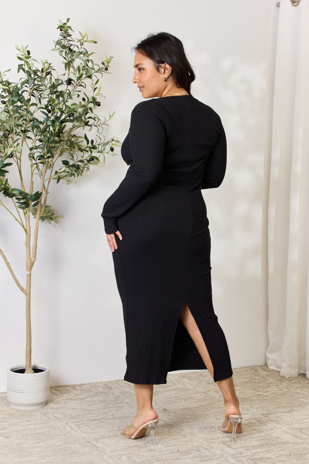 A person stands indoors wearing the chic Culture Code Full Size Ribbed Long Sleeve Midi Slit Dress paired with sandals, next to a potted plant.