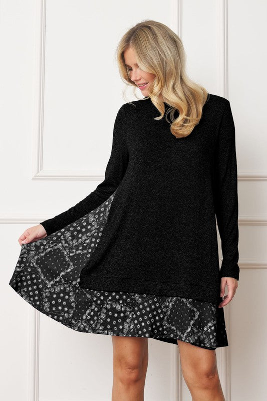 A woman with long blonde hair wears a grey long-sleeve sweater layered over the Mock Layered Bandana Paisley Mini Dress. She is standing indoors against a plain white wall and looking downward.