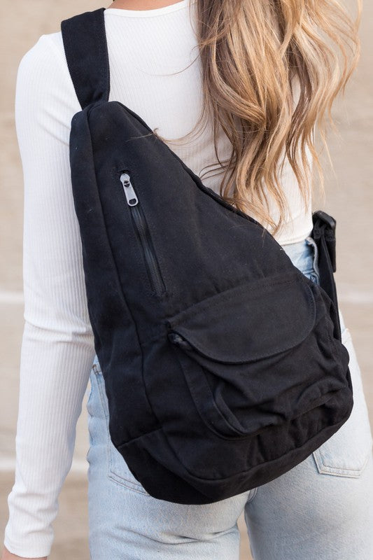 A person with long blonde hair carries the Oversized Canvas Sling, featuring an adjustable strap, while wearing a white long-sleeve top and light blue jeans. The image is taken from behind.