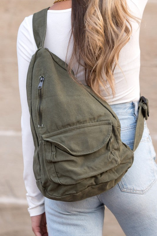 A person with long blonde hair carries the Oversized Canvas Sling, featuring an adjustable strap, while wearing a white long-sleeve top and light blue jeans. The image is taken from behind.