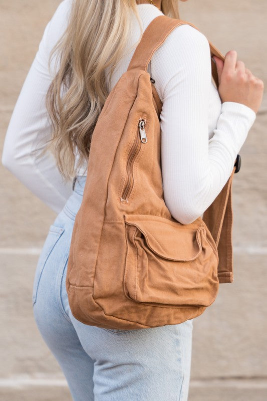 A person with long blonde hair carries the Oversized Canvas Sling, featuring an adjustable strap, while wearing a white long-sleeve top and light blue jeans. The image is taken from behind.