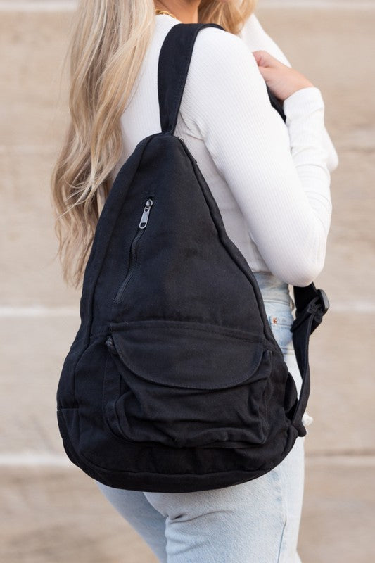 A person with long blonde hair carries the Oversized Canvas Sling, featuring an adjustable strap, while wearing a white long-sleeve top and light blue jeans. The image is taken from behind.