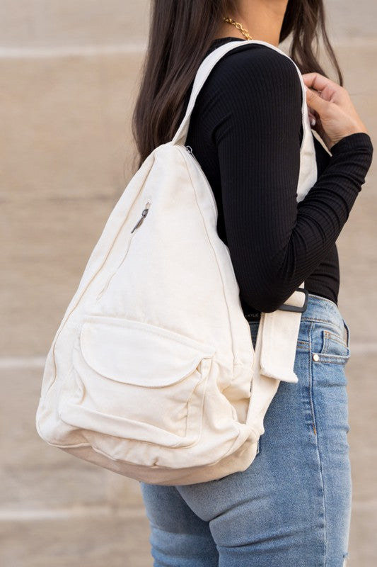 A person with long blonde hair carries the Oversized Canvas Sling, featuring an adjustable strap, while wearing a white long-sleeve top and light blue jeans. The image is taken from behind.