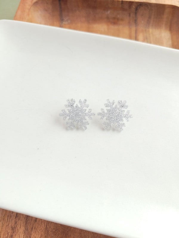A pair of Snowflake Studs, glittery and crafted from hypoallergenic stainless steel, displayed on a wooden surface.