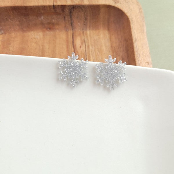 A pair of Snowflake Studs, glittery and crafted from hypoallergenic stainless steel, displayed on a wooden surface.