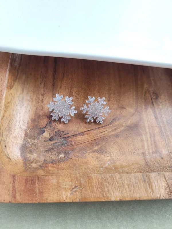 A pair of Snowflake Studs, glittery and crafted from hypoallergenic stainless steel, displayed on a wooden surface.