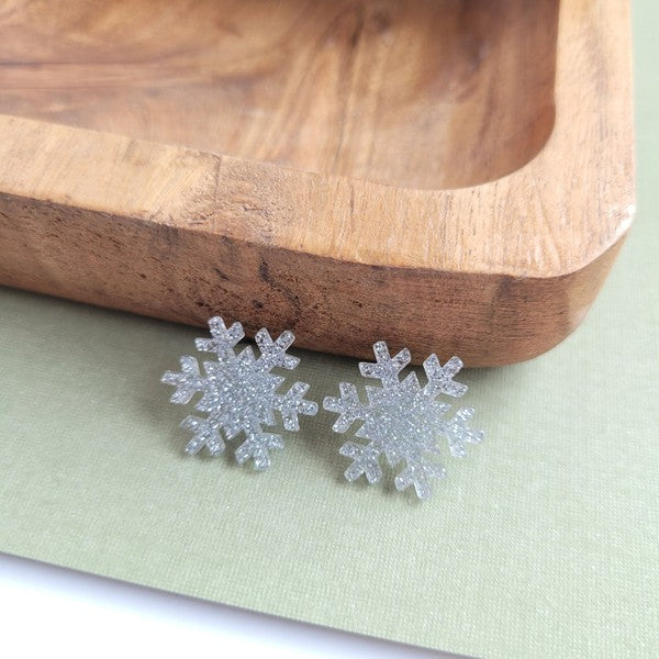 A pair of Snowflake Studs, glittery and crafted from hypoallergenic stainless steel, displayed on a wooden surface.