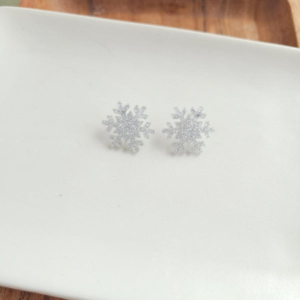 A pair of Snowflake Studs, glittery and crafted from hypoallergenic stainless steel, displayed on a wooden surface.