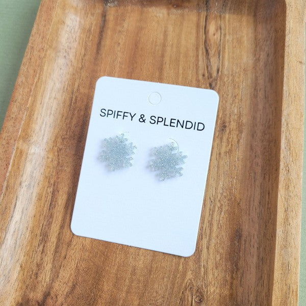 A pair of Snowflake Studs, glittery and crafted from hypoallergenic stainless steel, displayed on a wooden surface.