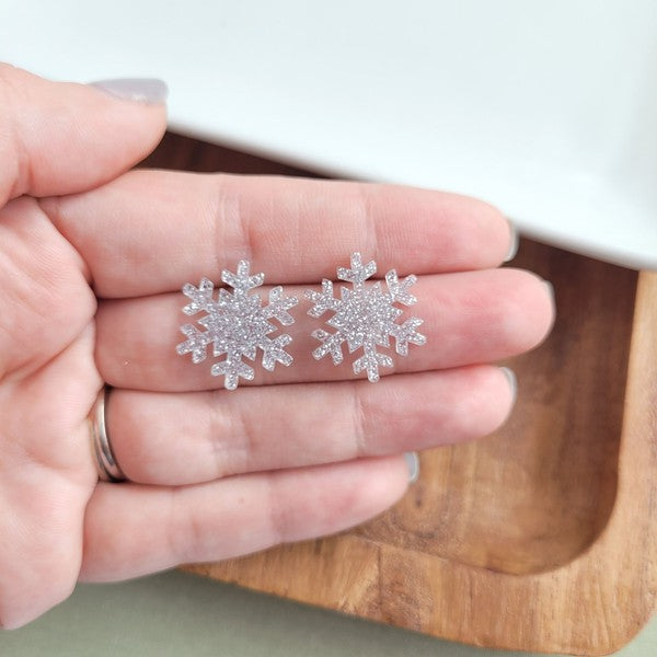 A pair of Snowflake Studs, glittery and crafted from hypoallergenic stainless steel, displayed on a wooden surface.