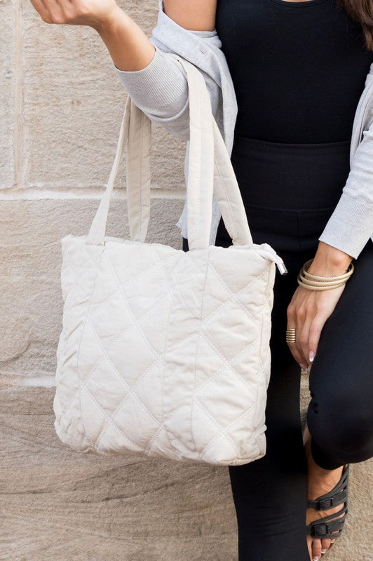 A person is holding the Quilted Tote, a grey bag with a zipper closure, in front of a stone wall.