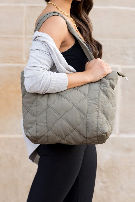 A person is holding the Quilted Tote, a grey bag with a zipper closure, in front of a stone wall.