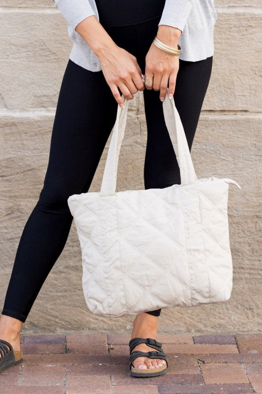 A person is holding the Quilted Tote, a grey bag with a zipper closure, in front of a stone wall.