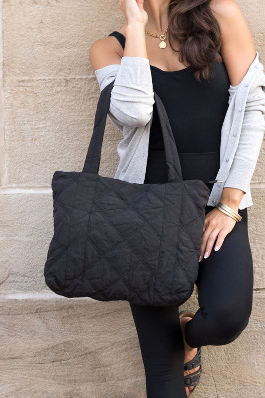 A person is holding the Quilted Tote, a grey bag with a zipper closure, in front of a stone wall.