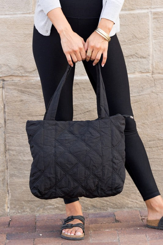 A person is holding the Quilted Tote, a grey bag with a zipper closure, in front of a stone wall.