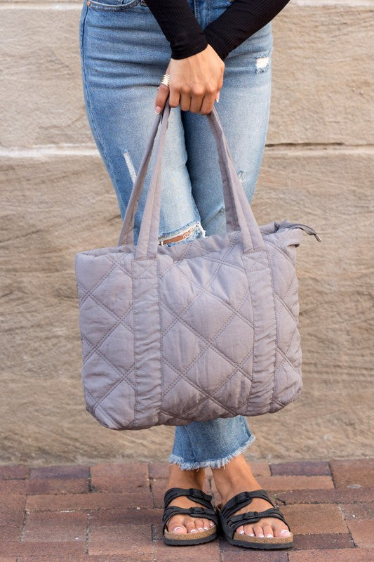 A person is holding the Quilted Tote, a grey bag with a zipper closure, in front of a stone wall.