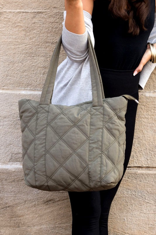 A person is holding the Quilted Tote, a grey bag with a zipper closure, in front of a stone wall.