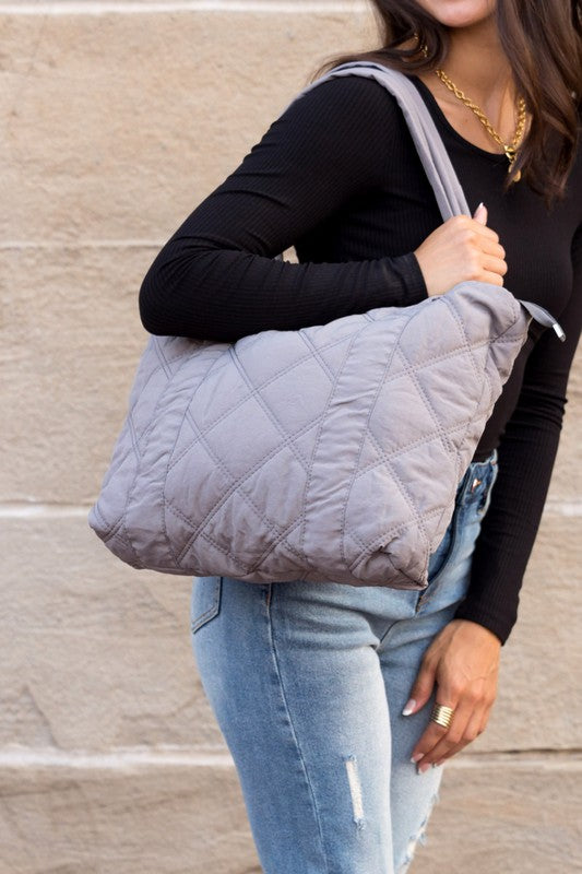 A person is holding the Quilted Tote, a grey bag with a zipper closure, in front of a stone wall.