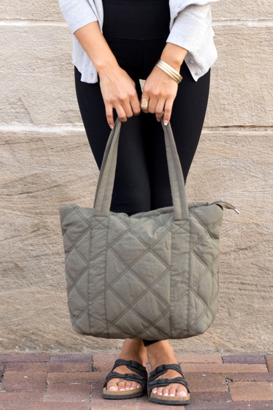 A person is holding the Quilted Tote, a grey bag with a zipper closure, in front of a stone wall.