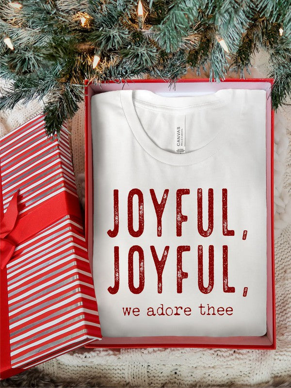 A Joyful, Joyful, We Adore Thee Short Sleeve Tee lies on a woven mat, featuring bold red text that reads "JOYFUL, JOYFUL, we adore thee" on the front.