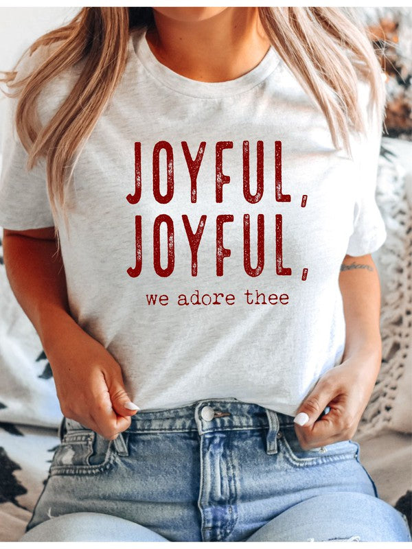 A Joyful, Joyful, We Adore Thee Short Sleeve Tee lies on a woven mat, featuring bold red text that reads "JOYFUL, JOYFUL, we adore thee" on the front.