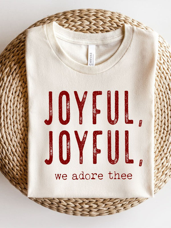 A Joyful, Joyful, We Adore Thee Short Sleeve Tee lies on a woven mat, featuring bold red text that reads "JOYFUL, JOYFUL, we adore thee" on the front.